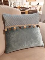 poduszka wykonana z najwyższej jakości welurowej tkaniny w odcieniu delikatnego błękitu, wpadającego w zieleń. Dodano także element z gładkiej tkaniny w kratkę. Dodatkowego uroku dodaje taśma z pomponami wszyta w poduszkę.